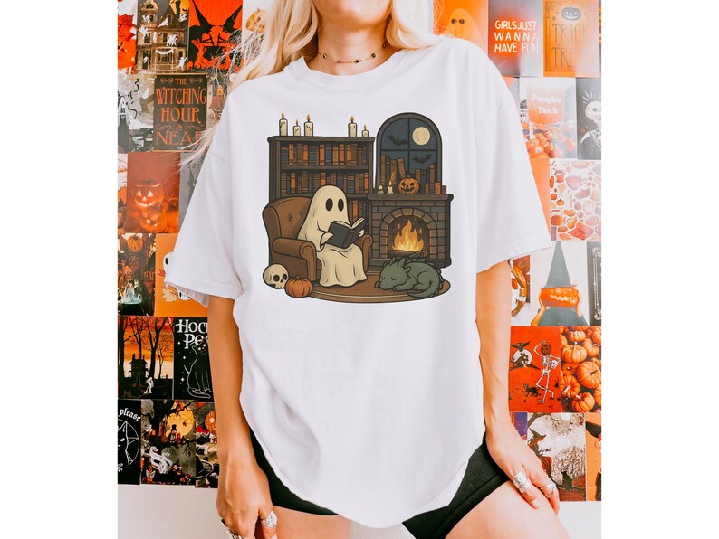 May include: White t-shirt featuring a cartoon ghost reading a book in front of a fireplace, with a cat, pumpkin, and skull. The scene is set in a cozy room with a bookshelf and a window with a moon. Halloween theme.