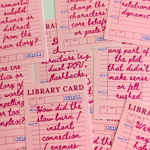 May include: Pink library cards with handwritten questions in red ink, such as "romantic subplot enhance or distract from the main story?" and "How did the slow burn / instant connection / enemies?"