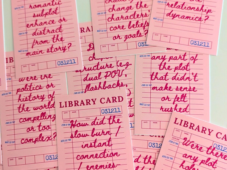 May include: Pink library cards with handwritten questions in red ink, such as "romantic subplot enhance or distract from the main story?" and "How did the slow burn / instant connection / enemies?"