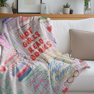 May include: A soft, printed throw blanket draped over a white sofa. The blanket features a collage of book-themed graphics, including the text "HOT GIRLS READ BOOKS" in red, and "ONE MORE CHAPTER" in blue. Pink hearts and book illustrations are also present.