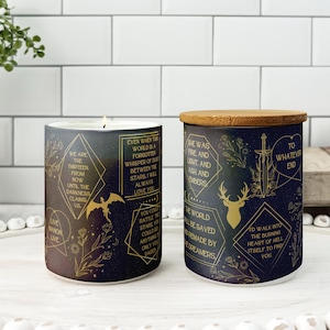 May include: Two decorative candles with dark blue and gold designs. One is lit, the other has a wooden lid. The candles feature text and illustrations of a dragon, deer, and sword. The candles are on a white surface.