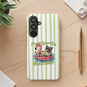May include: A phone case with a green and white striped background. The case features a raccoon wearing sunglasses and floating on a pink flamingo floatie, holding a drink. The text "Margarita Weather" is also on the case.