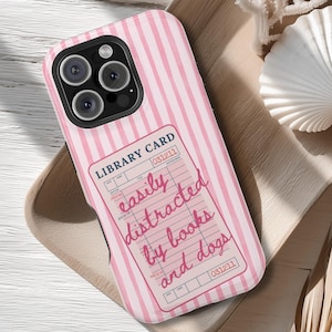 May include: Pink and white striped phone case with a library card design. The card reads "easily distracted by books and dogs" in a handwritten font. The case has a camera cutout and is on a wooden tray.
