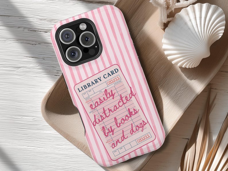 May include: Pink and white striped phone case with a library card design. The card reads "easily distracted by books and dogs" in a handwritten font. The case has a camera cutout and is on a wooden tray.