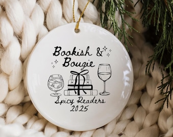 May include: White ceramic Christmas ornament with the text "Bookish & Bougie Spicy Readers 2025". The ornament features illustrations of a cocktail, books, and a wine glass, with a gold string for hanging.