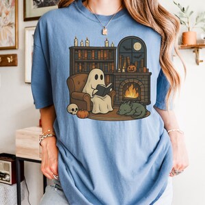 May include: Light blue t-shirt featuring a cartoon ghost reading a book by a fireplace. The illustration includes a bookshelf, pumpkin, skull, and a sleeping dragon. The design has a cozy, autumnal theme.