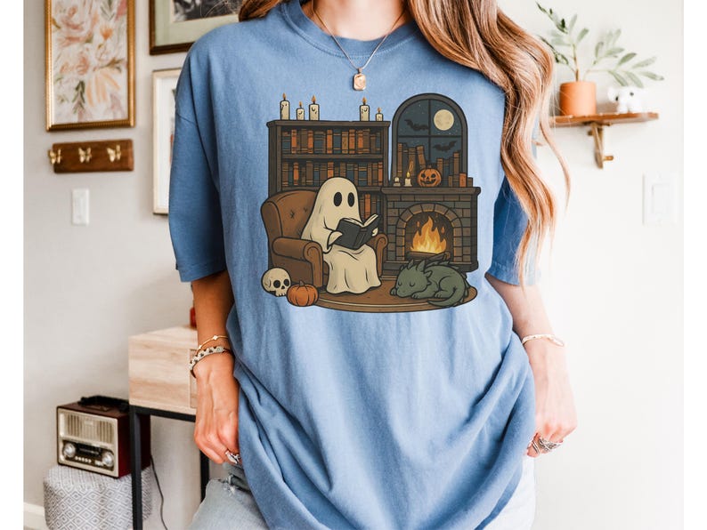May include: Light blue t-shirt featuring a cartoon ghost reading a book by a fireplace. The illustration includes a bookshelf, pumpkin, skull, and a sleeping dragon. The design has a cozy, autumnal theme.