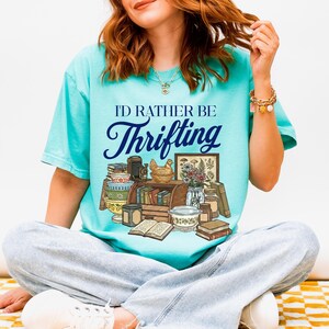 May include: A turquoise t-shirt with the text "I'd Rather Be Thrifting" in a script font. The graphic features a collection of vintage items, including books, a chicken figurine, and a framed botanical print. The person is wearing light blue jeans.
