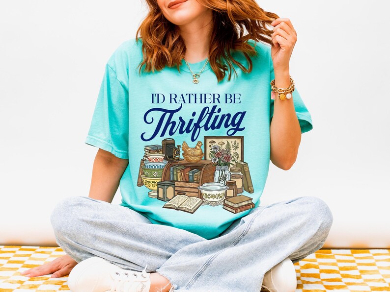 May include: A turquoise t-shirt with the text "I'd Rather Be Thrifting" in a script font. The graphic features a collection of vintage items, including books, a chicken figurine, and a framed botanical print. The person is wearing light blue jeans.