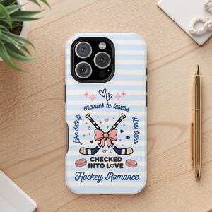 May include: A light blue and white striped phone case with a hockey romance theme. The case features crossed hockey sticks, a pink bow, and text that reads "enemies to lovers" and "checked into love".