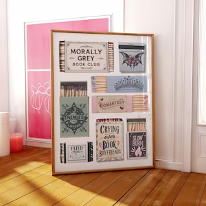 May include: A framed artwork featuring various matchbox designs. The matchboxes have different themes and text, including "Morally Grey Book Club," "Enemies to Lovers," and "Slow Burn." The artwork is displayed on a wooden floor near a window.