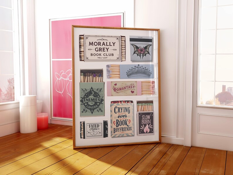 May include: A framed artwork featuring various matchbox designs. The matchboxes have different themes and text, including "Morally Grey Book Club," "Enemies to Lovers," and "Slow Burn." The artwork is displayed on a wooden floor near a window.