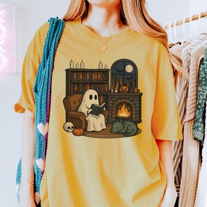 May include: Mustard yellow t-shirt featuring a cartoon ghost reading a book in a cozy setting. The design includes a fireplace, bookshelves, a sleeping dragon, a skull, and a pumpkin, creating a Halloween theme.