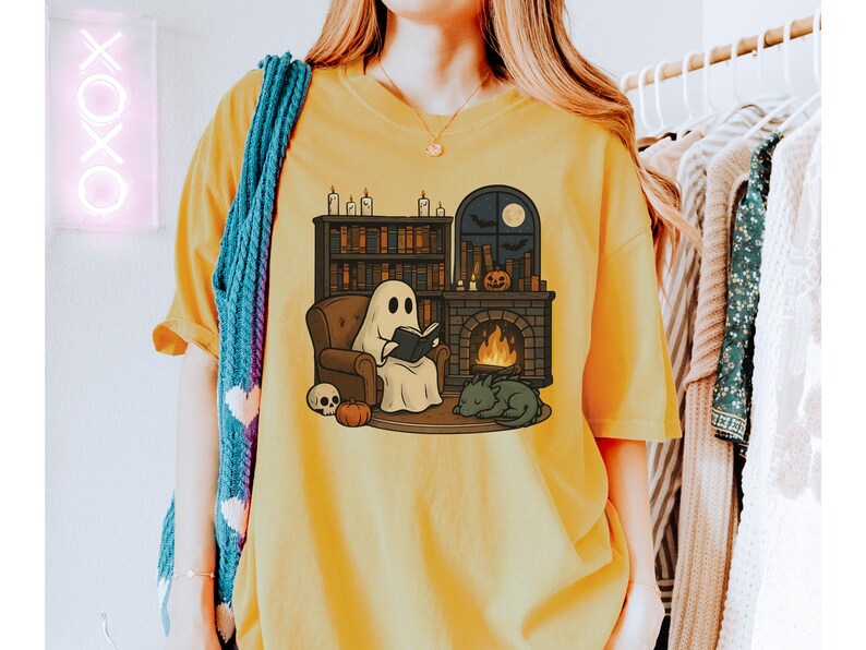 May include: Mustard yellow t-shirt featuring a cartoon ghost reading a book in a cozy setting. The design includes a fireplace, bookshelves, a sleeping dragon, a skull, and a pumpkin, creating a Halloween theme.