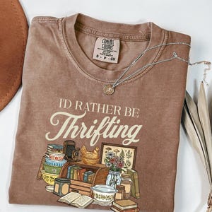 May include: Brown t-shirt with the phrase "I'D RATHER BE Thrifting" in a vintage font. The graphic includes illustrations of books, a camera, a chicken figurine, and other thrifted items. A silver necklace with a pendant is draped on the shirt.