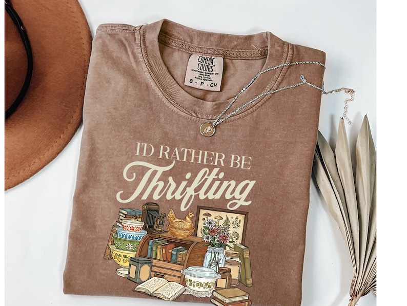 May include: Brown t-shirt with the phrase "I'D RATHER BE Thrifting" in a vintage font. The graphic includes illustrations of books, a camera, a chicken figurine, and other thrifted items. A silver necklace with a pendant is draped on the shirt.