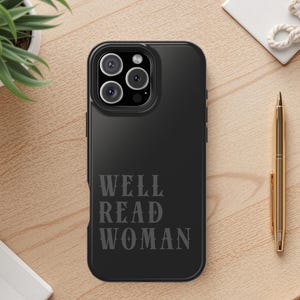 May include: Black phone case with the text "WELL READ WOMAN" in a serif font. The phone case has a triple-lens camera. A gold pen, a small white plant, and a white notepad are also visible.