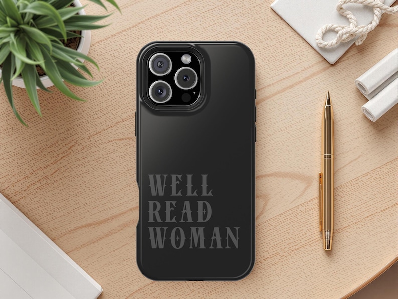 May include: Black phone case with the text "WELL READ WOMAN" in a serif font. The phone case has a triple-lens camera. A gold pen, a small white plant, and a white notepad are also visible.