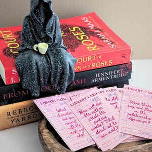 May include: A stack of books with red, black, and tan covers, topped with a dark figurine holding a small yellow cup. Pink library cards with handwritten questions are in a wooden bowl.