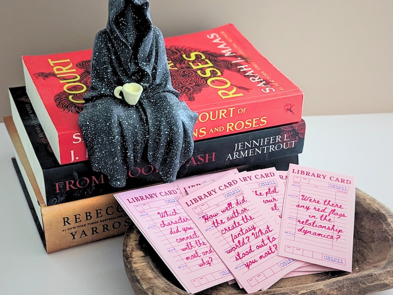 May include: A stack of books with red, black, and tan covers, topped with a dark figurine holding a small yellow cup. Pink library cards with handwritten questions are in a wooden bowl.