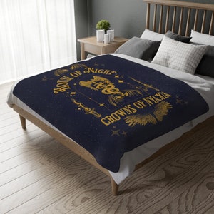 May include: A navy blue fleece blanket with a gold design featuring a winged serpent, a dagger, and the text "House of Night Crowns of Nyaxia".