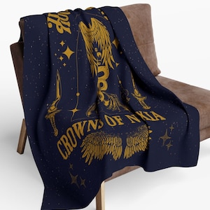 May include: A navy blue throw blanket with a gold design featuring a winged serpent, two daggers, and the text "Crowns of Nyxia". The design is set against a starry background.