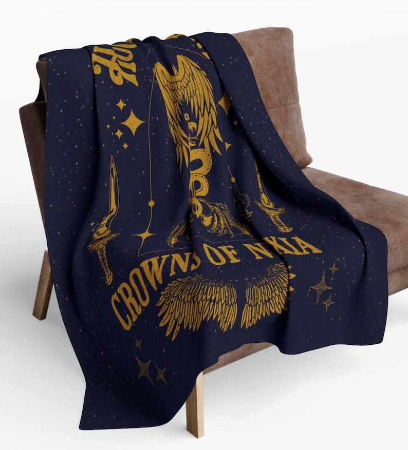 May include: A navy blue throw blanket with a gold design featuring a winged serpent, two daggers, and the text "Crowns of Nyxia". The design is set against a starry background.
