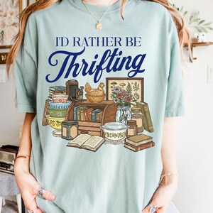 May include: A light blue t-shirt with the text "I'D RATHER BE Thrifting" in navy blue script. The graphic features a collection of vintage items, including books, bowls, a camera, and a framed picture.