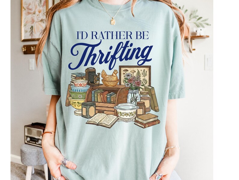 May include: A light blue t-shirt with the text "I'D RATHER BE Thrifting" in navy blue script. The graphic features a collection of vintage items, including books, bowls, a camera, and a framed picture.
