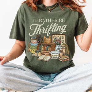 May include: Olive green t-shirt with the text "I'd Rather Be Thrifting" above a graphic of vintage items. The graphic includes books, bowls, a chicken figurine, and a framed botanical print. The shirt is a relaxed fit.