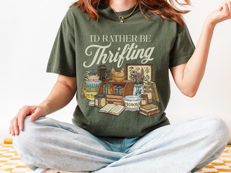 May include: Olive green t-shirt with the text "I'd Rather Be Thrifting" above a graphic of vintage items. The graphic includes books, bowls, a chicken figurine, and a framed botanical print. The shirt is a relaxed fit.