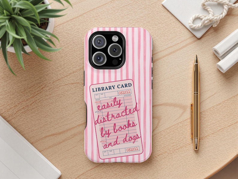 May include: Pink and white striped phone case with a library card design. The card reads "easily distracted by books and dogs" in a handwritten style. The phone case is on a wooden surface with a plant and a gold pen.