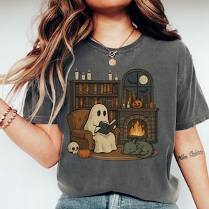 May include: Dark gray t-shirt featuring a cozy Halloween scene. A ghost reads a book in a chair by a fireplace, with a pumpkin, skull, and sleeping dragon. A bookshelf, candles, and a window with bats and a moon complete the design.