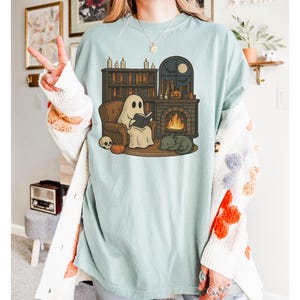 May include: Light blue t-shirt featuring a cartoon ghost reading a book in front of a fireplace, with a cat, pumpkin, and skull. The design includes a bookshelf, candles, and a moon. The shirt is a relaxed fit.