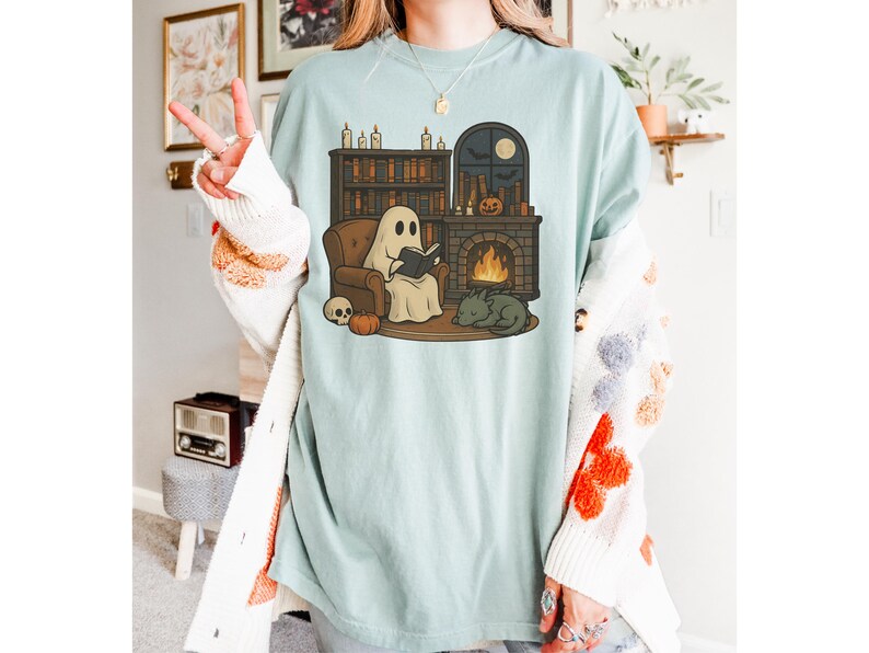 May include: Light blue t-shirt featuring a cartoon ghost reading a book in front of a fireplace, with a cat, pumpkin, and skull. The design includes a bookshelf, candles, and a moon. The shirt is a relaxed fit.