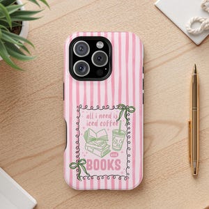 May include: Pink and white striped phone case with the text "all I need is iced coffee and books" in a decorative frame. The case has a camera cutout and is on a wooden surface.