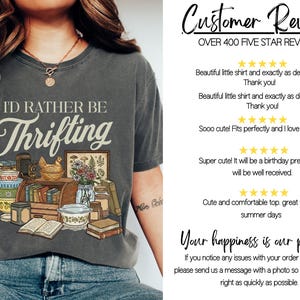 May include: Dark gray t-shirt with the text "I'd Rather Be Thrifting" in a vintage style. The graphic includes books, a camera, a chicken figurine, and a framed botanical print. The shirt is worn by a person.