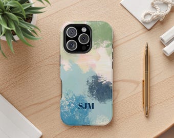 May include: A mobile phone case with an abstract watercolour design in green, blue, and pink hues. The case displays the initials "SJM" in navy blue. A gold pen and a small plant are also visible.