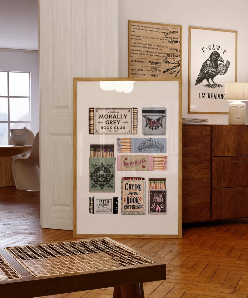 May include: A framed print featuring matchboxes with various designs and text, including "Morally Grey Book Club," "Enemies Lovers," and "Crying Over Book Boyfriends." The artwork is displayed in a room with wooden floors and a side table.