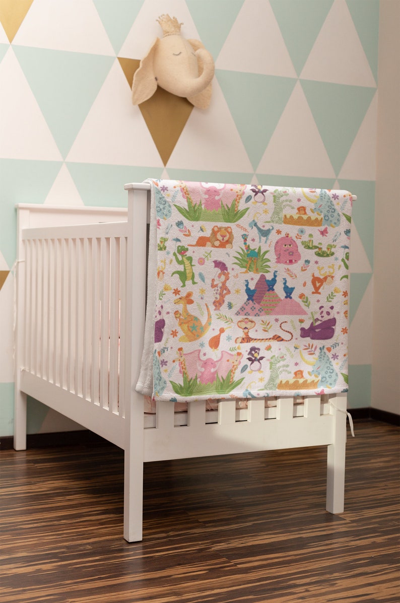 May include: White crib with a colorful animal print blanket hanging on the side. The blanket features a variety of animals, including a lion, a penguin, a kangaroo, and a giraffe. The crib is in a room with a light blue and white geometric wallpaper.