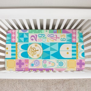 Op de afbeelding: Een hoeslaken voor een babybedje met een kleurrijk geometrisch patroon met een fantasierijk kasteelontwerp. Het laken is geel, blauw, roze, groen en goud met een herhalend patroon van geometrische vormen en cijfers.