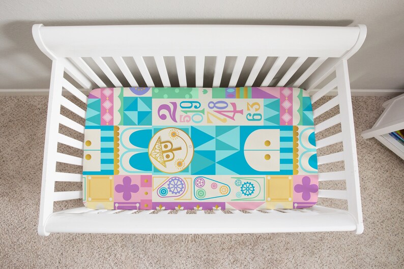 Op de afbeelding: Een hoeslaken voor een babybedje met een kleurrijk geometrisch patroon met een fantasierijk kasteelontwerp. Het laken is geel, blauw, roze, groen en goud met een herhalend patroon van geometrische vormen en cijfers.