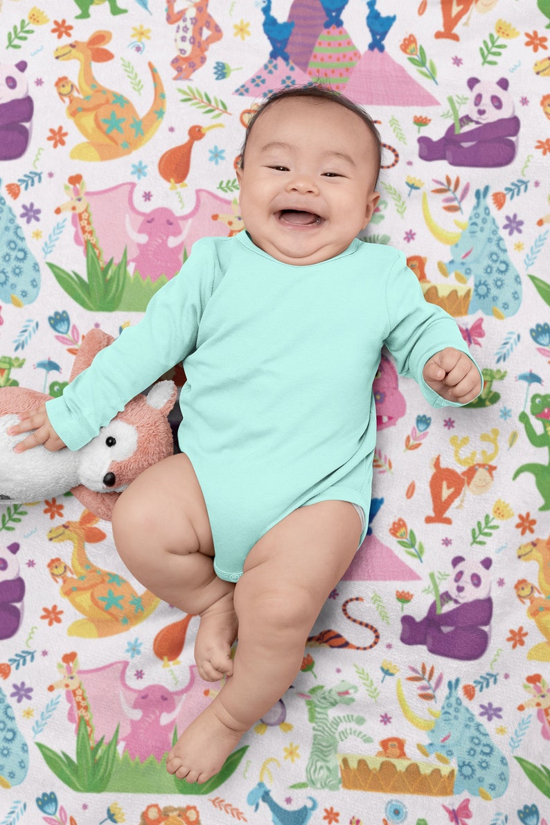 May include: A baby wearing a light blue long-sleeved onesie is lying on a white blanket with a colorful animal print. The baby is smiling and looking at the camera.