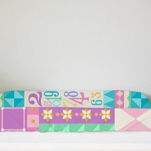 Op de afbeelding: Een kleurrijke, patchwork-stijl baby-verschoontafel met een geometrisch patroon met cijfers en bloemen. De tafel is gemaakt van zachte, comfortabele stof en is perfect voor het verschonen van baby's.