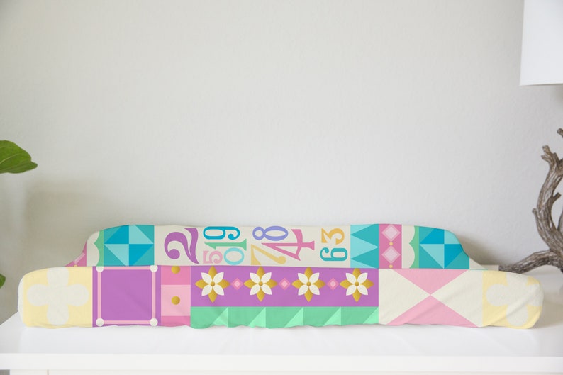 Op de afbeelding: Een kleurrijke, patchwork-stijl baby-verschoontafel met een geometrisch patroon met cijfers en bloemen. De tafel is gemaakt van zachte, comfortabele stof en is perfect voor het verschonen van baby's.