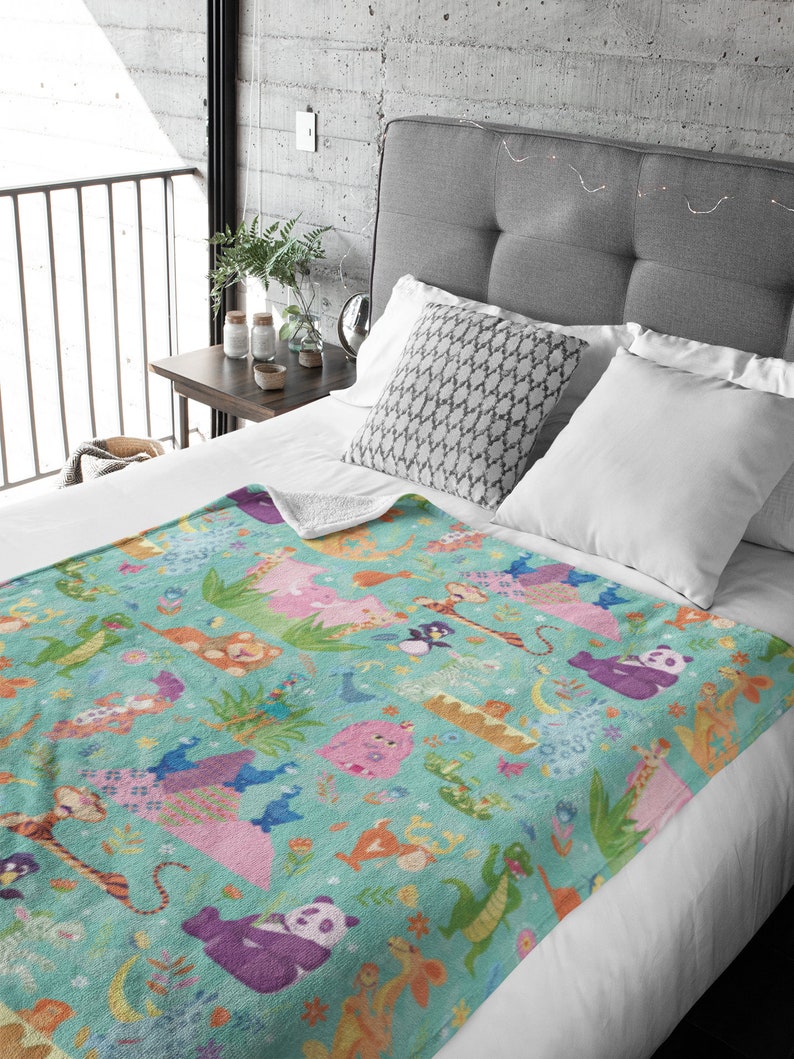 May include: A teal blue fleece blanket with a colorful pattern of animals, including a panda, a tiger, a monkey, and a dragon. The blanket is on a bed with white pillows and a gray headboard.