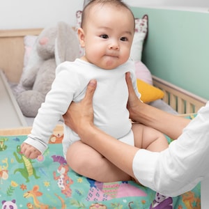 May include: A baby wearing a white onesie is being held by a person. The baby is sitting on a colorful blanket with a variety of animal illustrations. The blanket features a green background with a variety of animals including a panda, a tiger, a kangaroo, a kiwi, and a penguin.