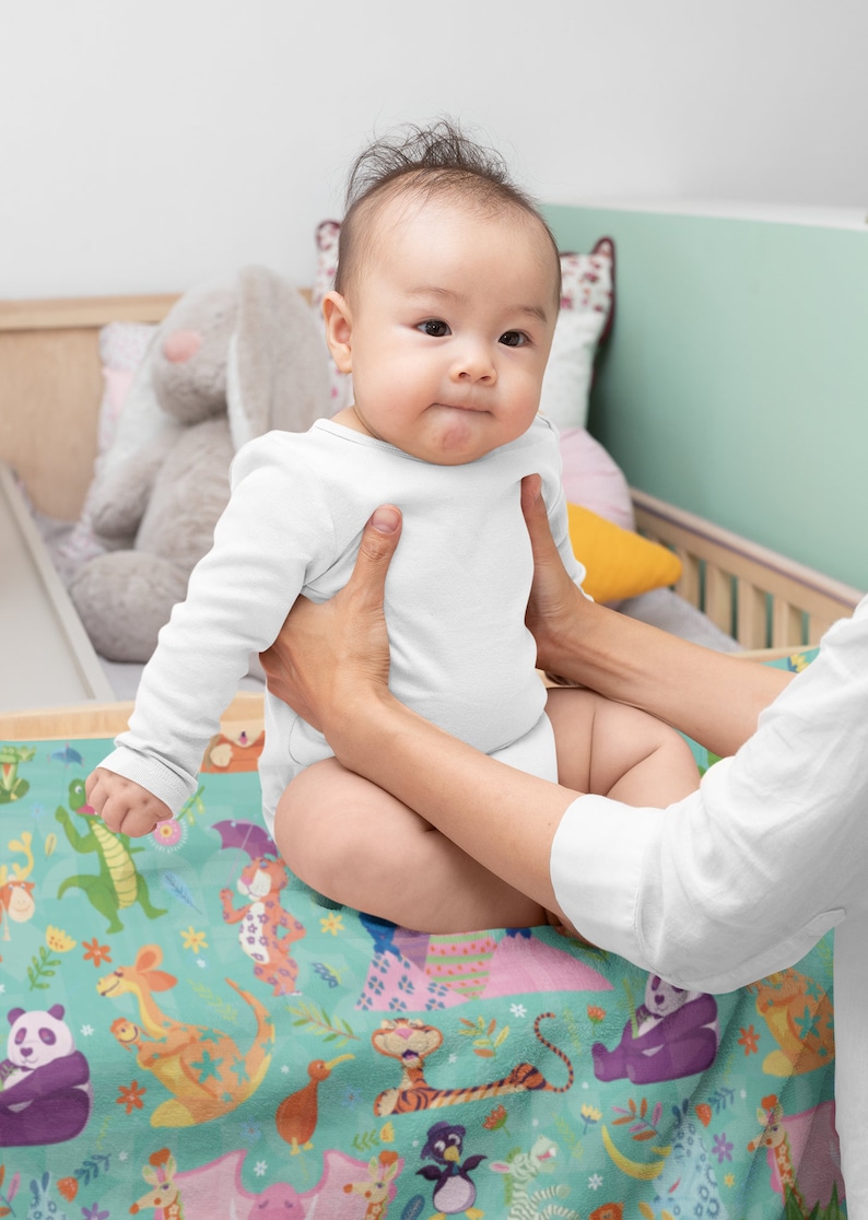 May include: A baby wearing a white onesie is being held by a person. The baby is sitting on a colorful blanket with a variety of animal illustrations. The blanket features a green background with a variety of animals including a panda, a tiger, a kangaroo, a kiwi, and a penguin.