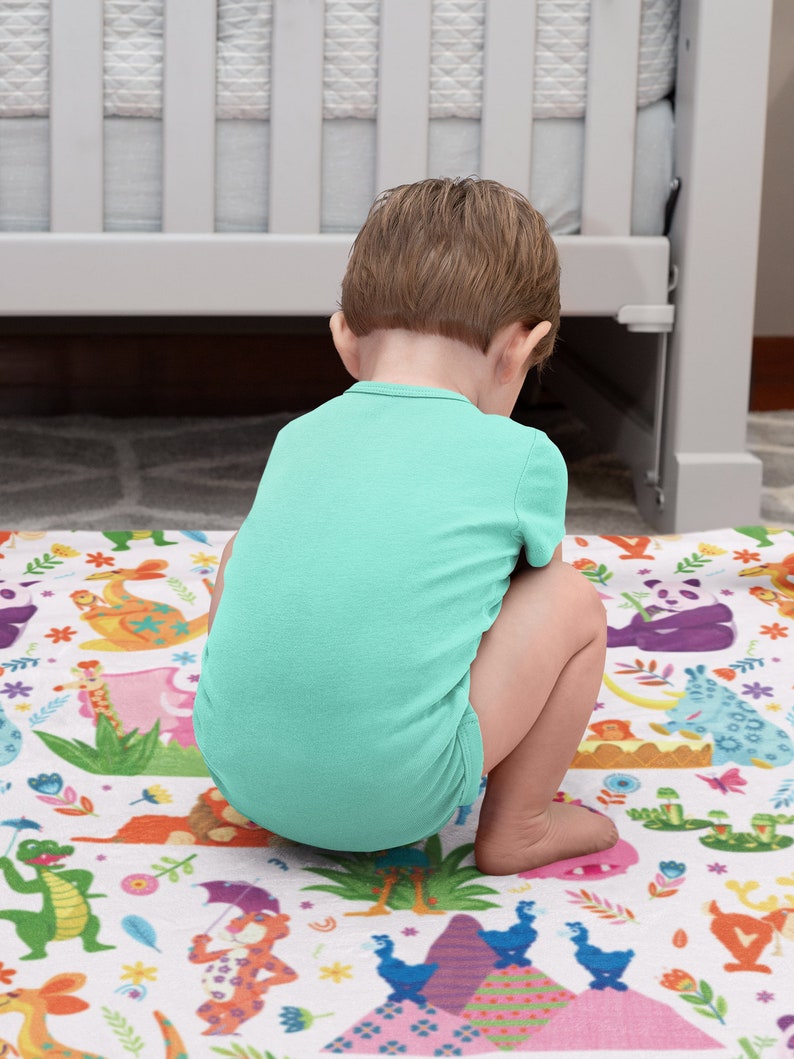 May include: A young child sits on a colorful play mat with a variety of animal illustrations. The child is wearing a light blue shirt and is looking down at the mat.