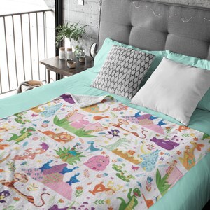 May include: A white fleece blanket with a colorful pattern of cartoon animals and flowers. The blanket is on a bed with a light blue sheet and gray headboard.